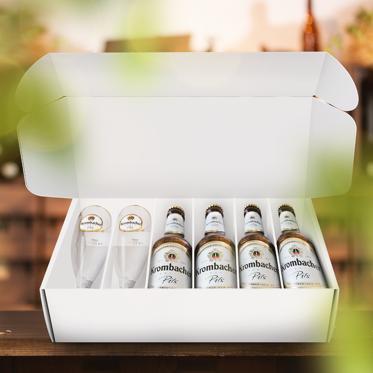  Weiße geöffnete Geschenkbox mit vier Pilsflaschen und zwei Gläsern darin.