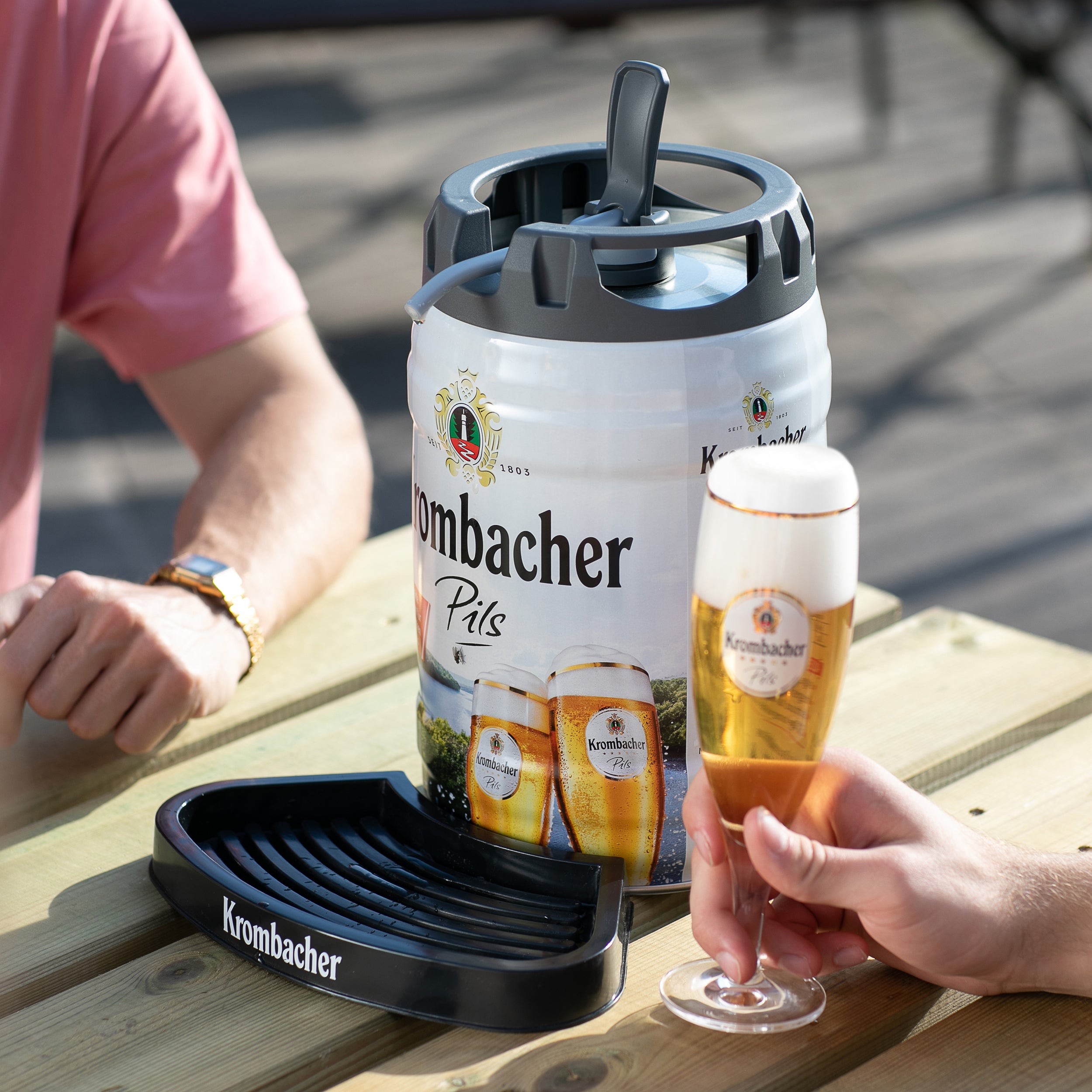 Krombacher-Pils-Fass mit Tropfschale auf Holztisch, eine Hand hält ein gefülltes Krombacher-Glas, eine Person mit rosa Hemd sitzt daneben