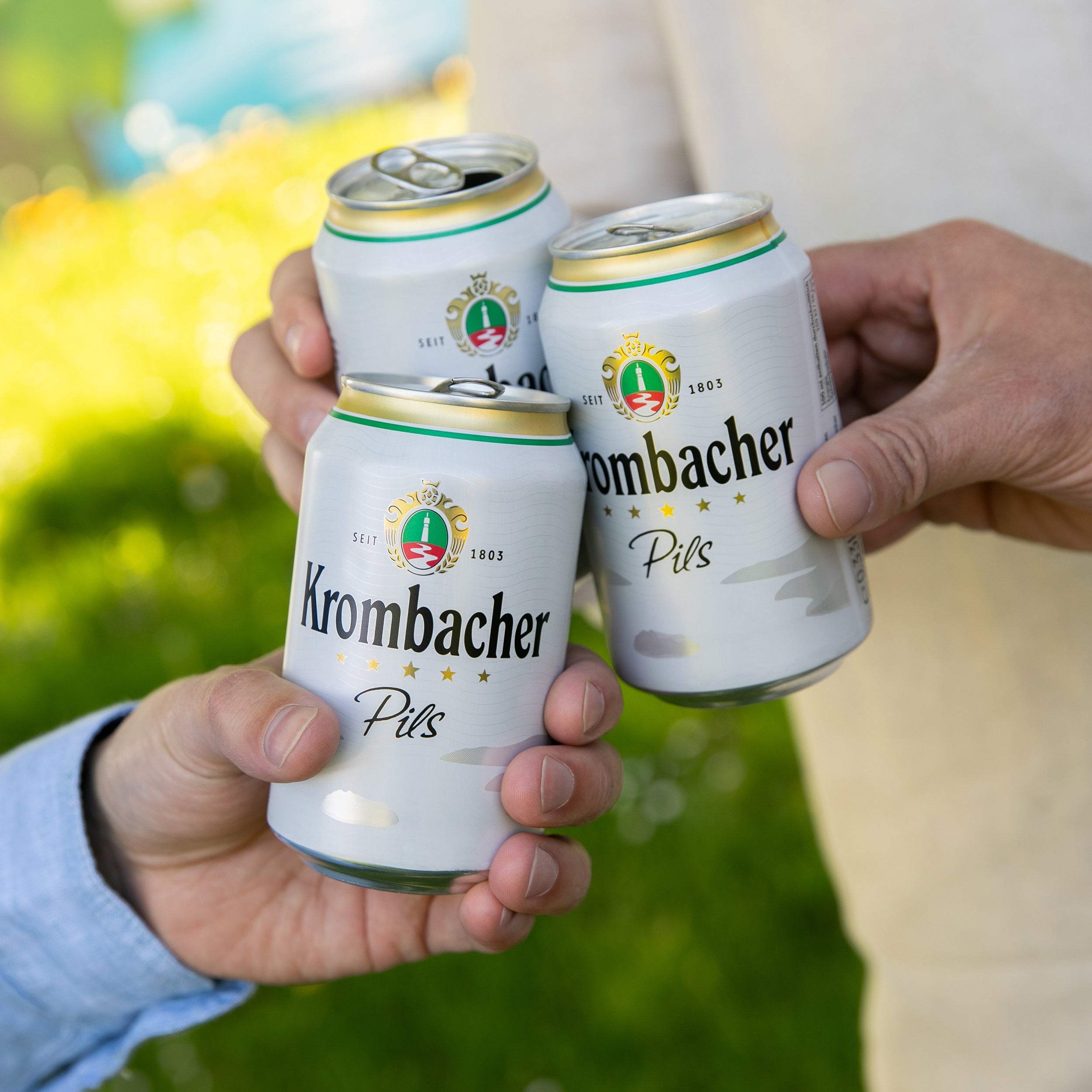 Drei Personen stoßen mit Krombacher 0,33l Dosen in der Natur an.