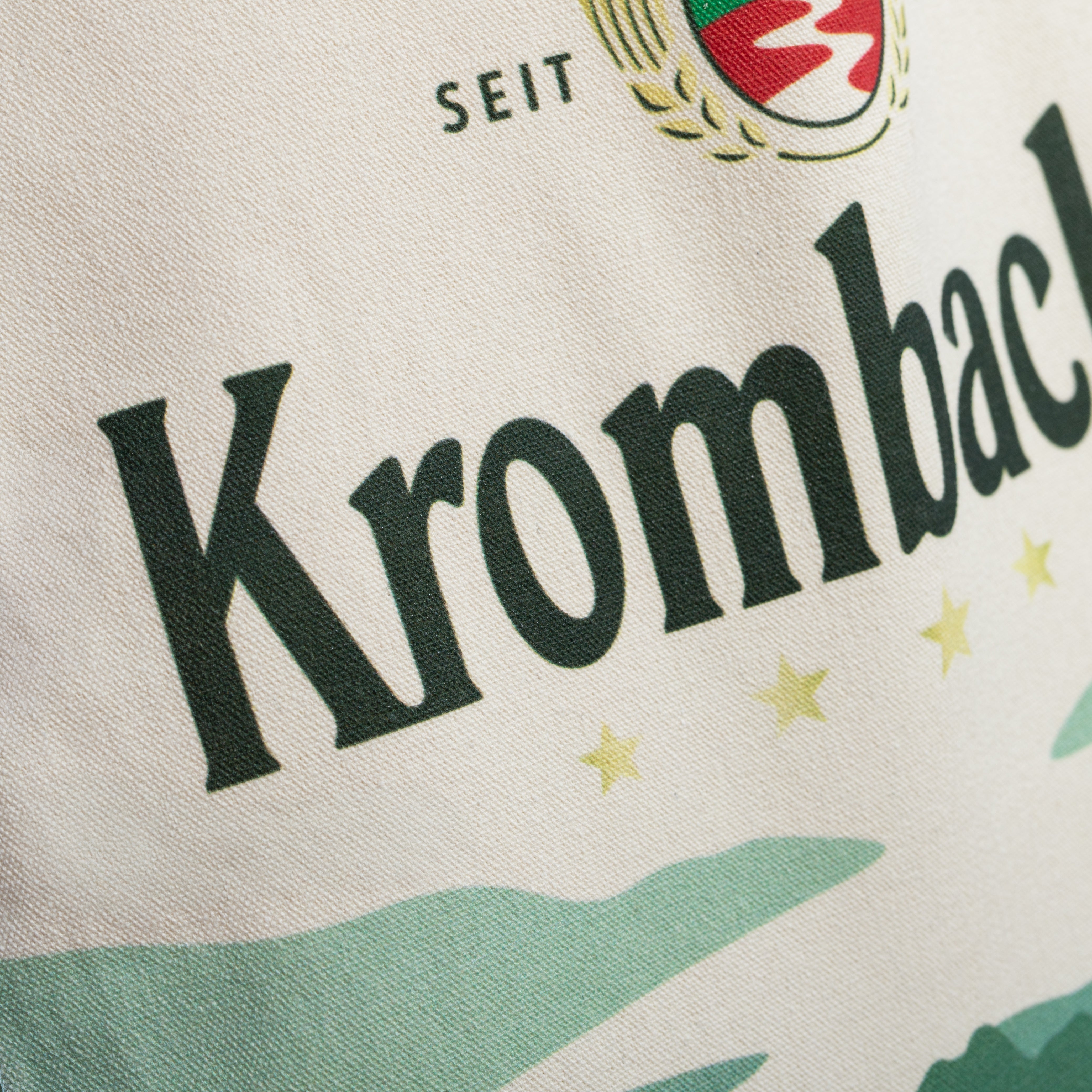 Nahaufnahme eines hellen Jutebeutels mit dem schwarzen Schriftzug 'Krombacher' und dem Teil eines Logos mit roten und goldenen Elementen.