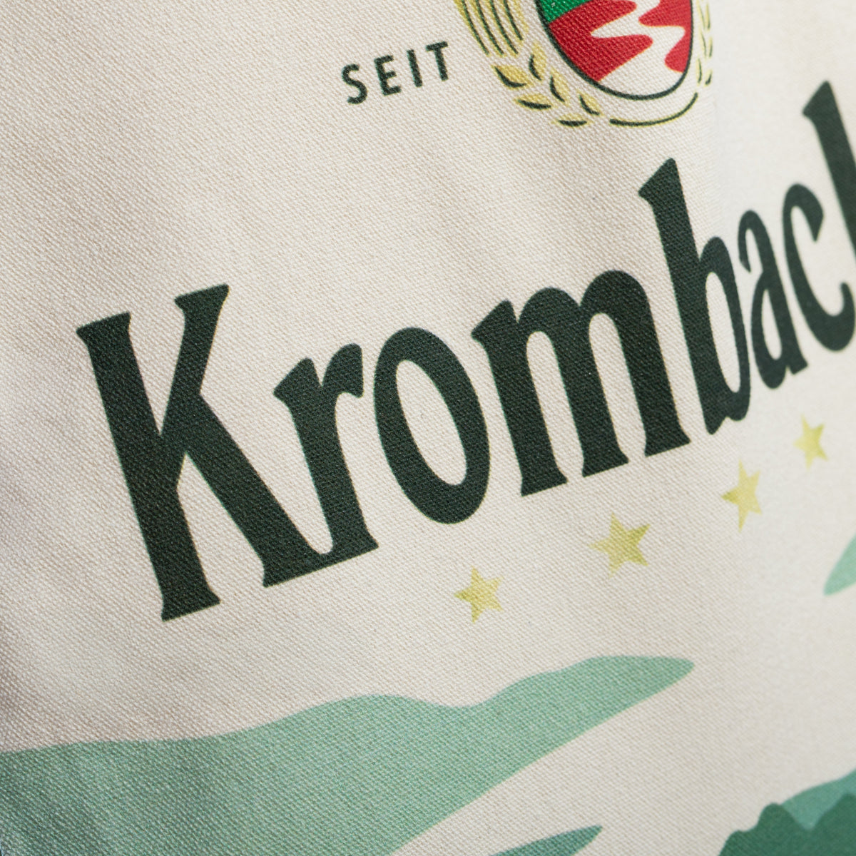 Nahaufnahme eines hellen Jutebeutels mit dem schwarzen Schriftzug 'Krombacher' und dem Teil eines Logos mit roten und goldenen Elementen.