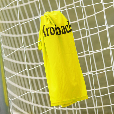 Gelbes Markierungshemd mit schwarzem Schriftzug 'Krombacher' hängt am Fußballtornetz.