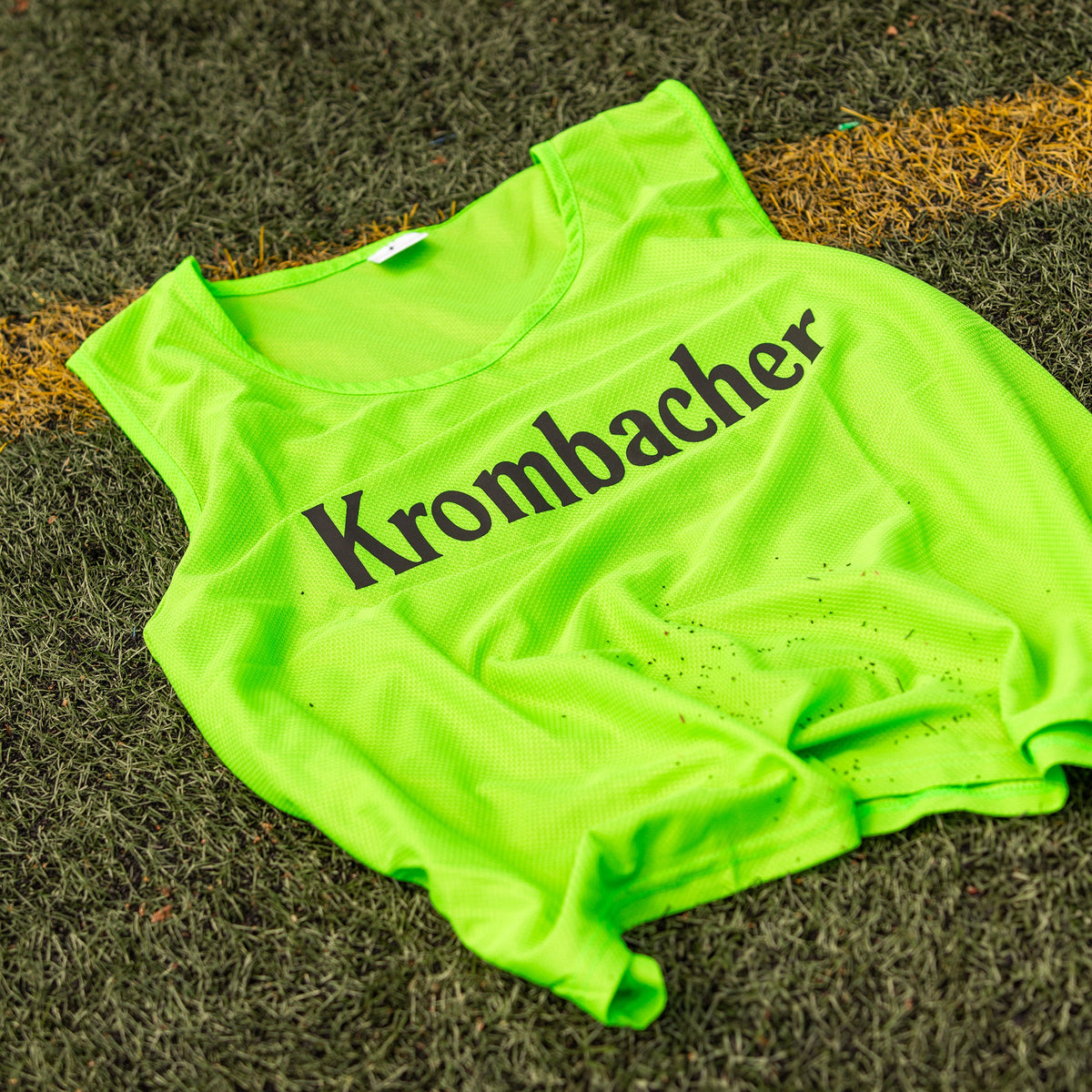 Grünes Markierungshemd mit schwarzem Schriftzug 'Krombacher' liegt auf dem rasen.