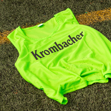 Grünes Markierungshemd mit schwarzem Schriftzug 'Krombacher' liegt auf dem rasen.