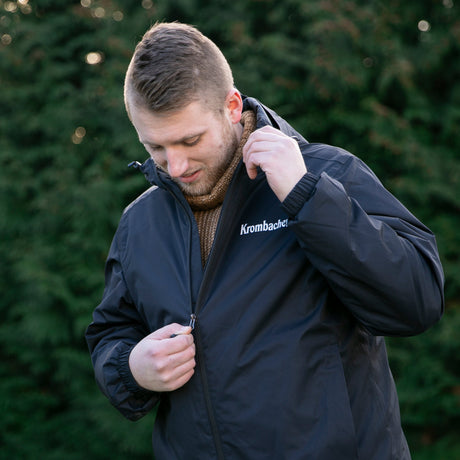 Person zieht schwarze Krombacher Regenjacke mit Reißverschluss und Kapuze an, trägt braunen Pullover darunter.