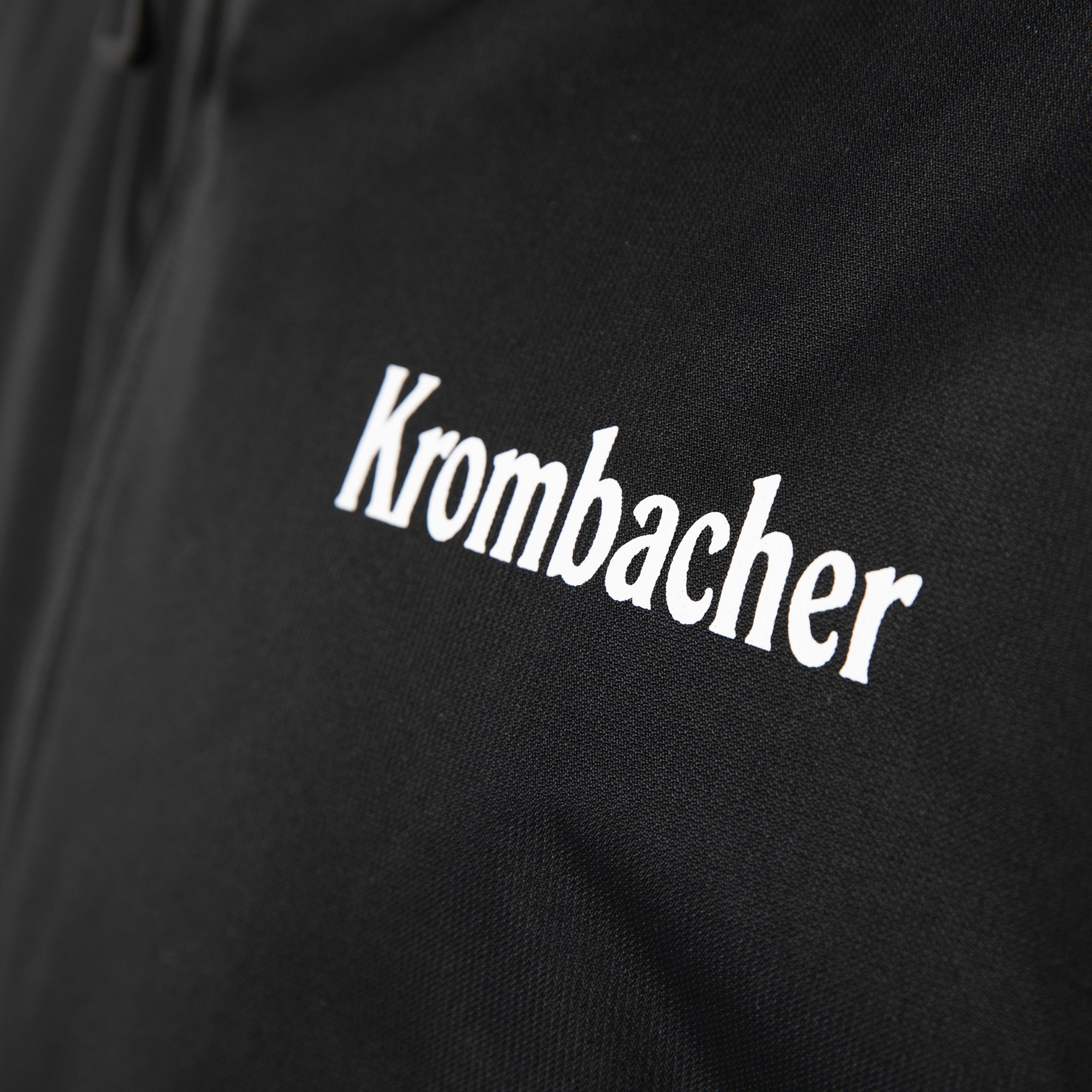 Schwarze Regenjacke mit weißem Schriftzug 'Krombacher' auf der Brust.