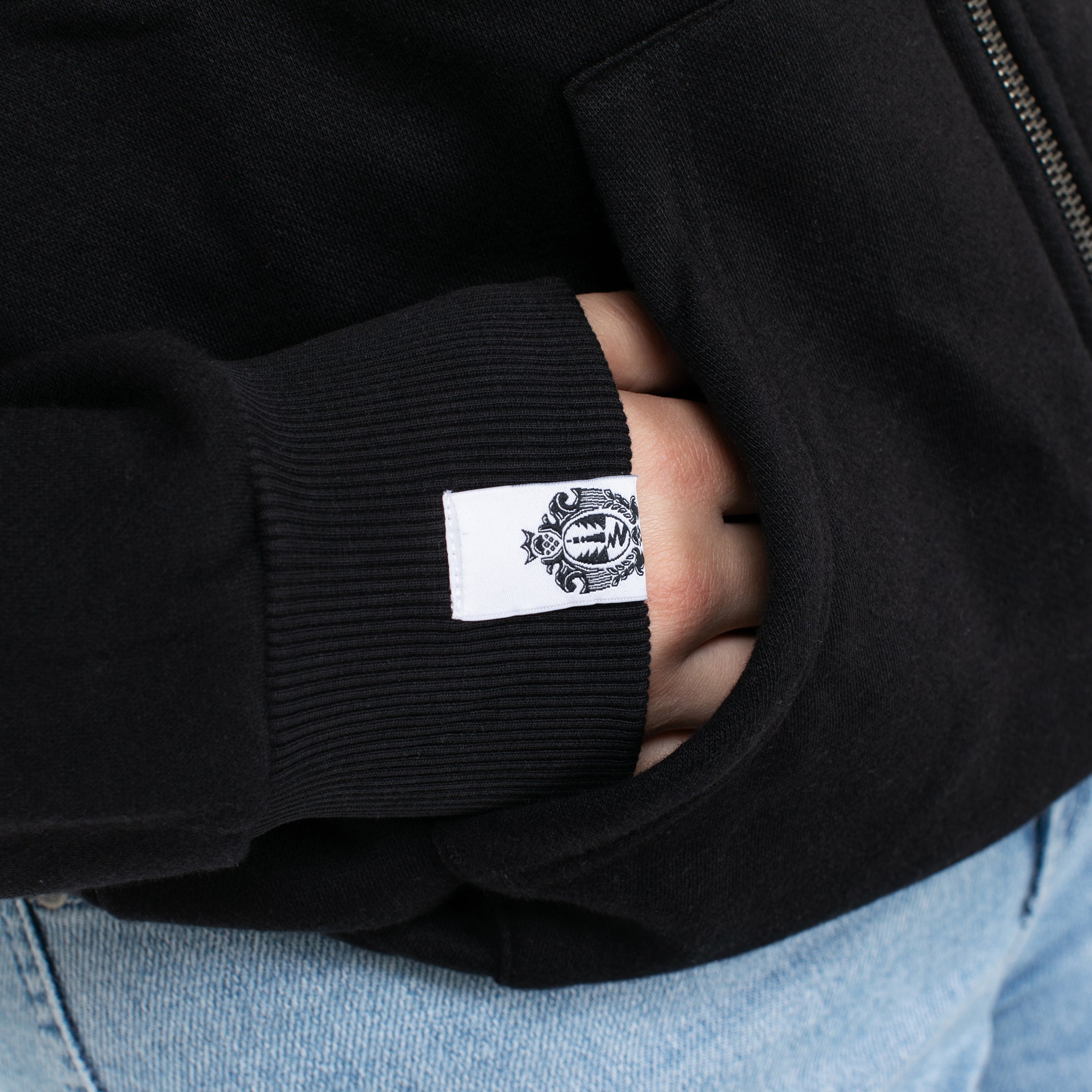 Hand steckt in der Tasche einer schwarzen Sweatshirtjacke mit weißem Krombacher-Logo auf dem Ärmelbündchen.