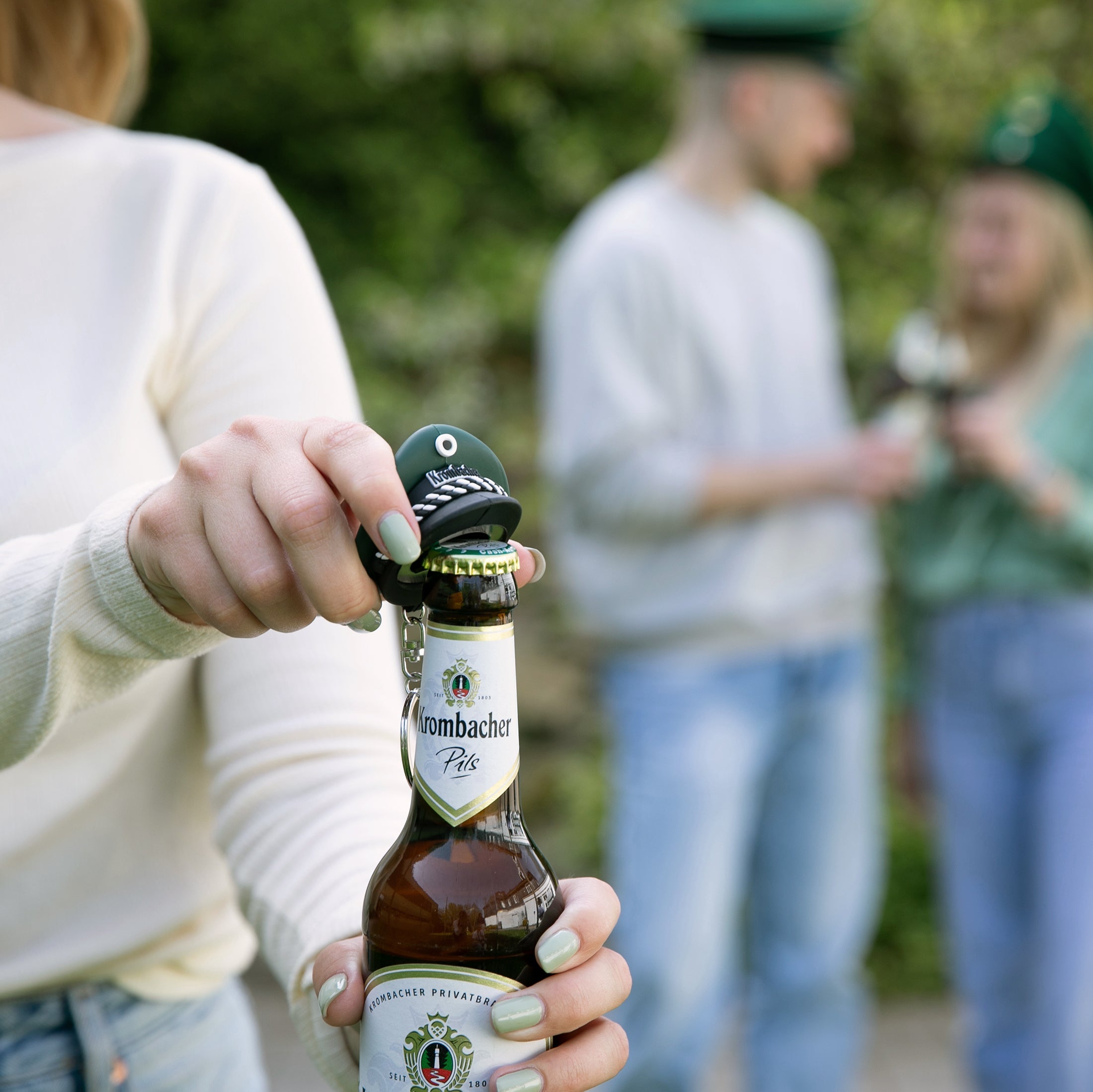 Hand öffnet Krombacher Pils Bierflasche mit Schlüsselanhänger in Form einer grünen Schützenkappe.