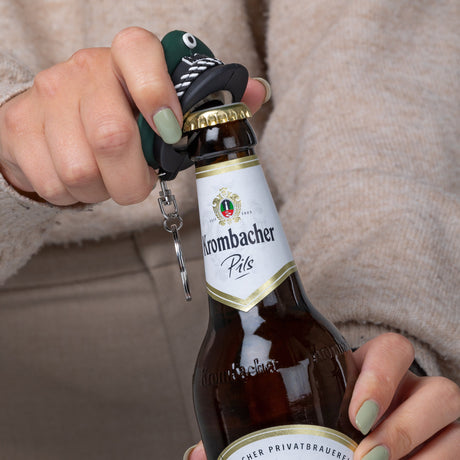 Hand öffnet Krombacher Pils Flasche mit schwarzem Flaschenöffner in Form einer Schützenkappe.