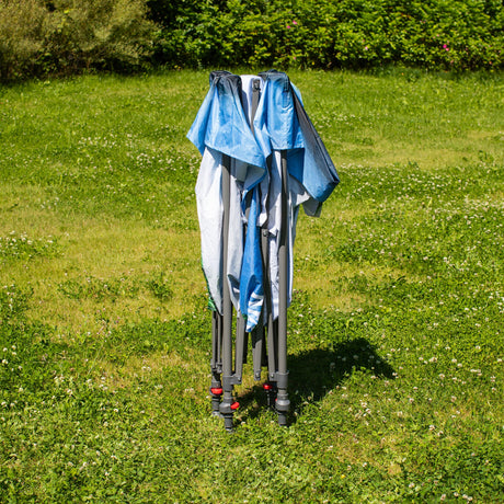 Zusammengeklappter blauer und weißer Pavillon auf einer grünen Wiese mit Büschen im Hintergrund.