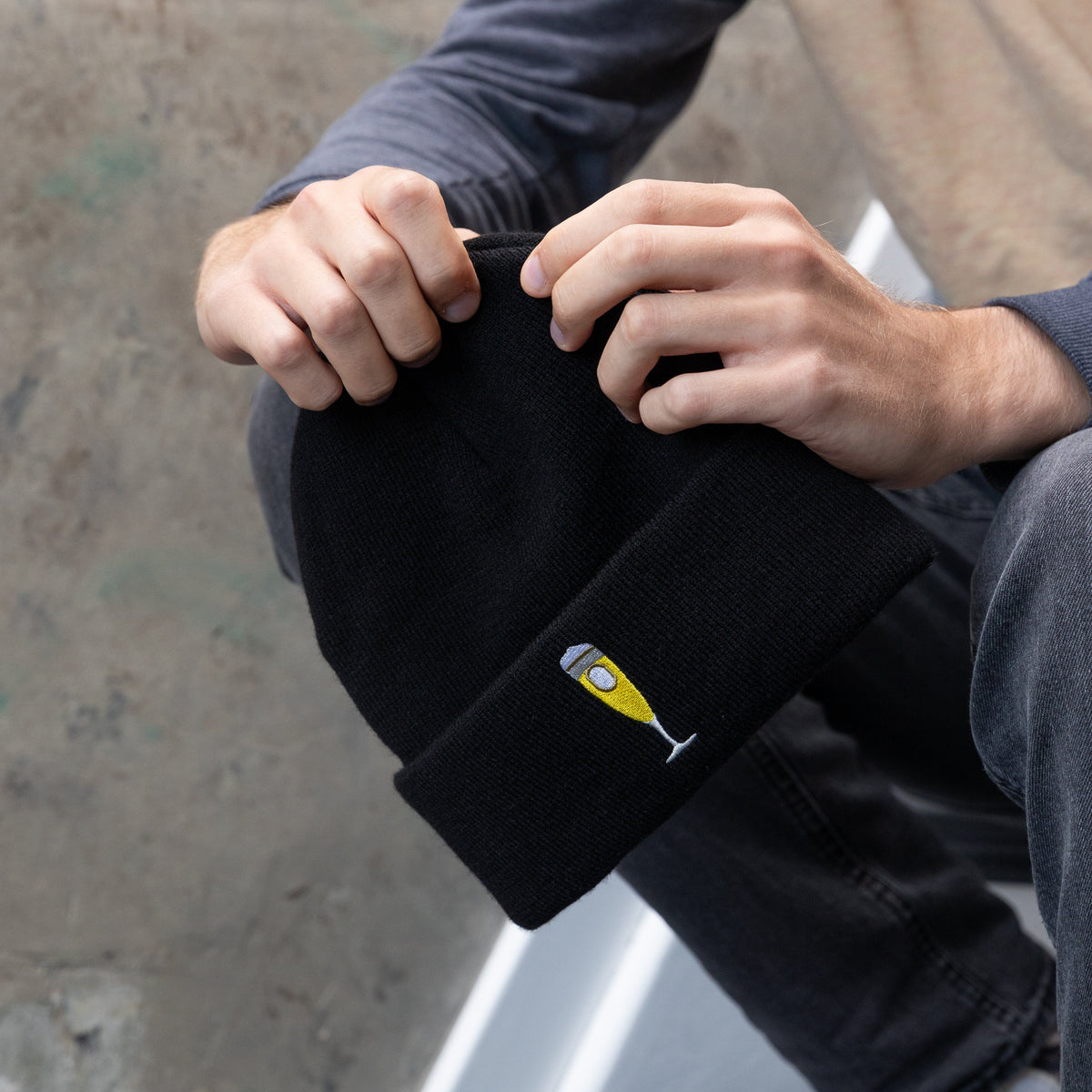 Person hält schwarze Beanie mit gesticktem Bierglas-Logo in der Hand.