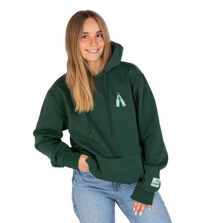 Frau trägt dunkelgrünen Hoodie mit kleinem Flaschendruck auf der Brust und Jeans, eine Hand in der Hosentasche