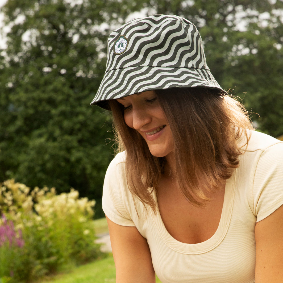 Frau mit wellenförmig gemustertem Bucket Hat und hellem T-Shirt sitzt lächelnd im Freien