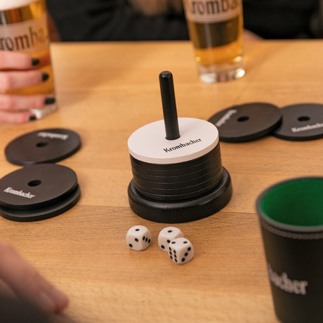 Krombacher Schockbesteck Spielscheiben mit schwarzer und weißer Farbe und Krombacher Schriftzug, Würfel und zwei Biergläser auf Holztisch.