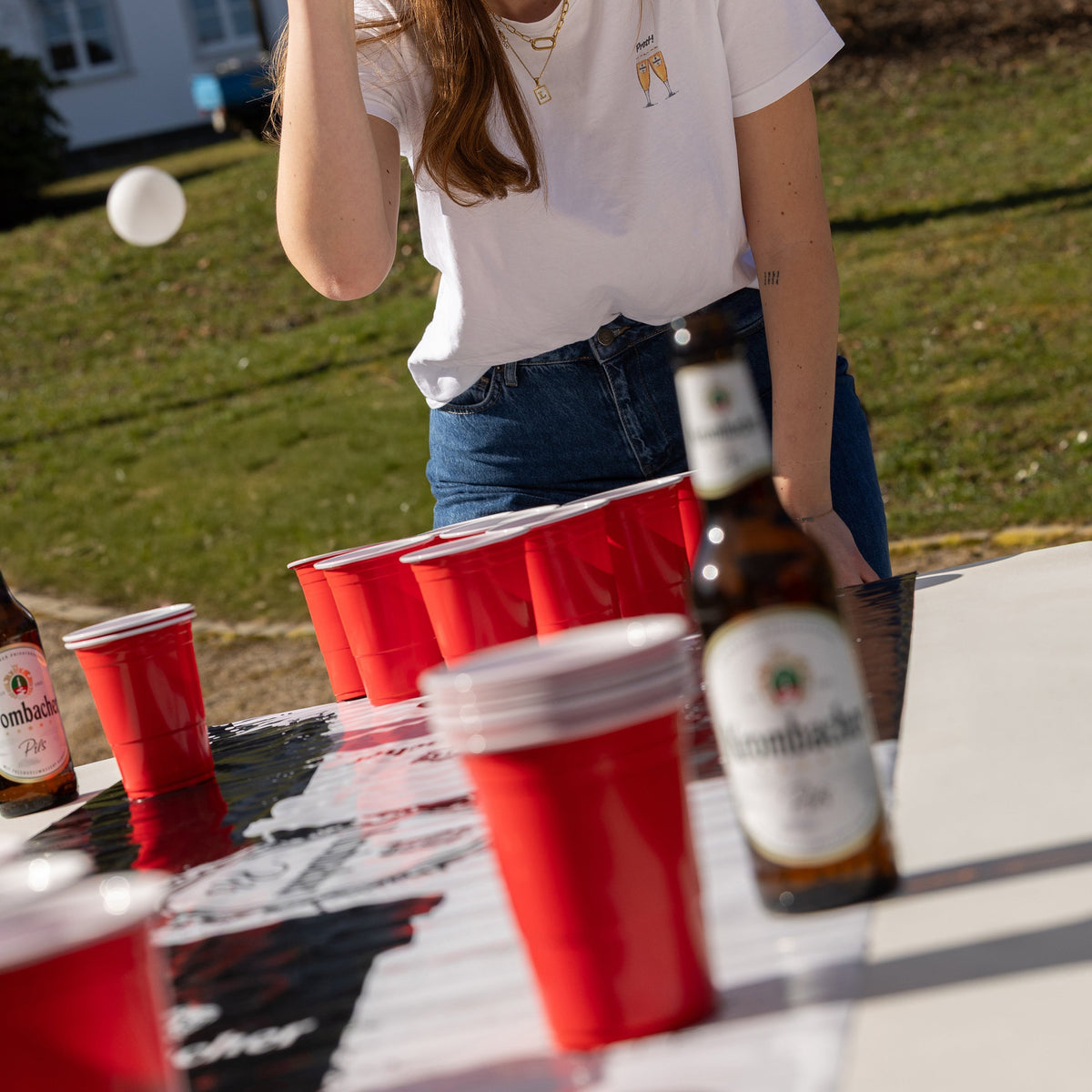 Person spielt Beerpong mit roten Bechern auf Krombacher-Matte auf weißem Tisch im Freien, Krombacher Pils Flasche daneben.