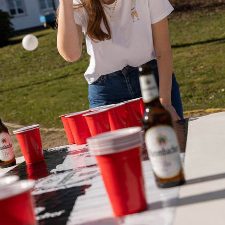 Person spielt Beerpong mit roten Bechern auf Krombacher-Matte auf weißem Tisch im Freien, Krombacher Pils Flasche daneben.