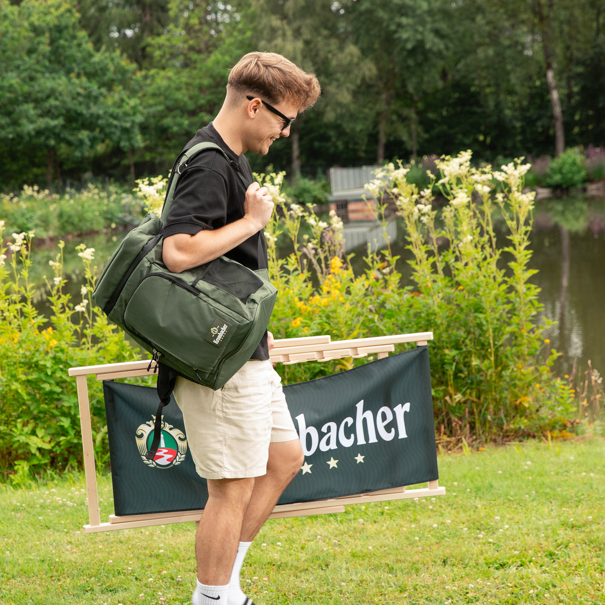 Junger Mann mit Sonnenbrille trägt zusammengeklappten Krombacher Liegestuhl und grüne Tasche in Parkanlage neben Teich