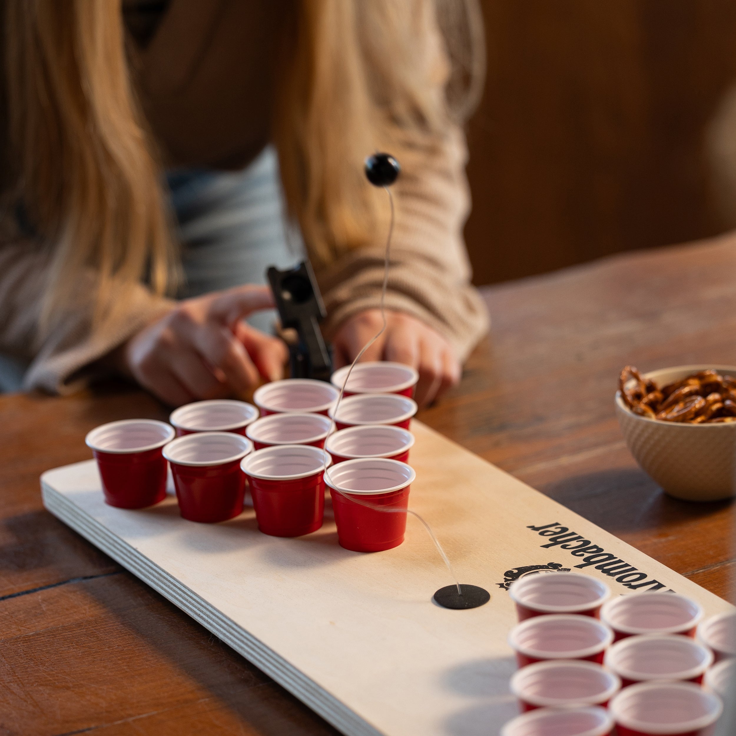 Person spielt Mini Beer Pong mit roten Bechern auf einem Krombacher-Brett, schwarzer Ball an Schnur in der Luft.