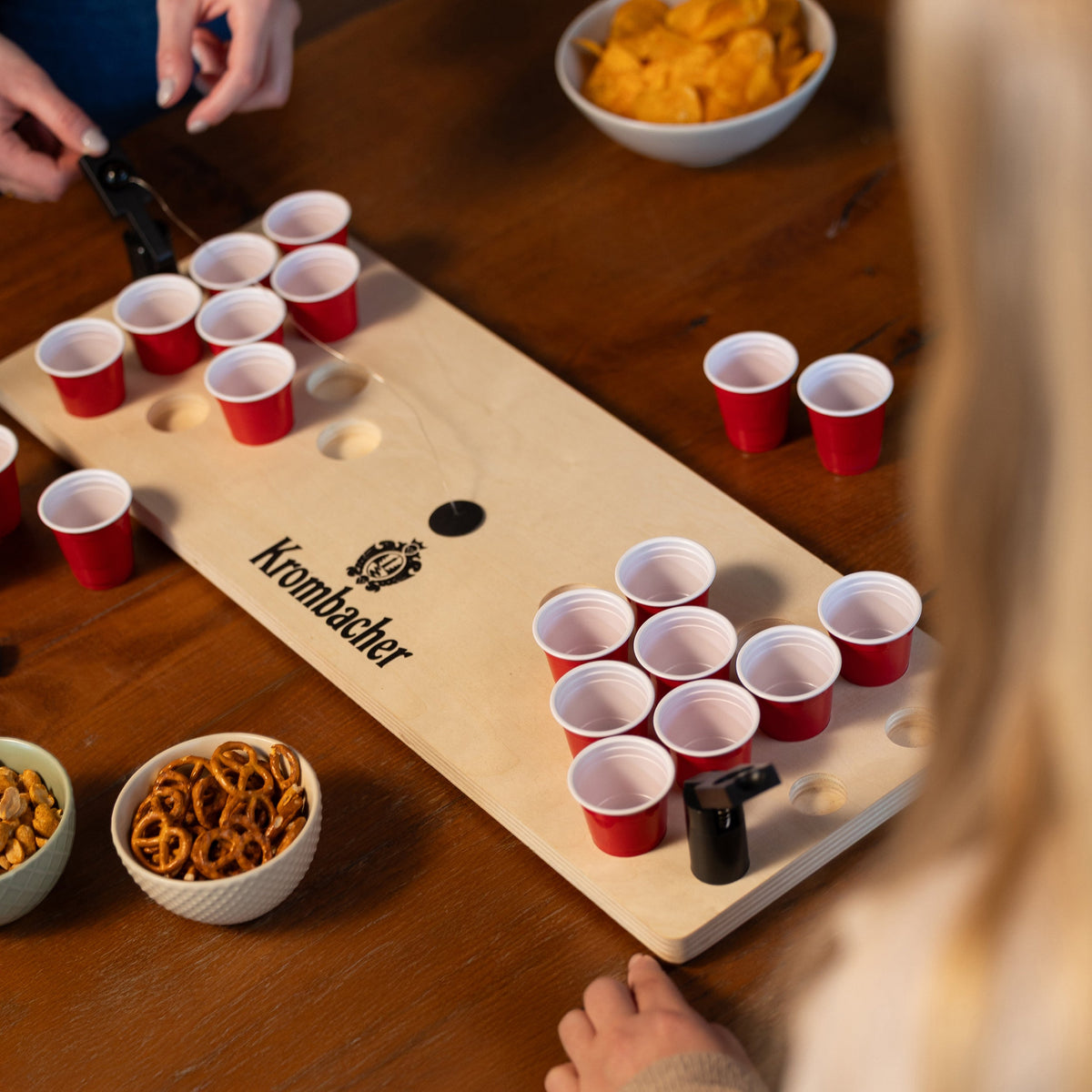 Mini Beerpong-Spielbrett mit roten Bechern und Krombacher-Logo, umgeben von Snacks wie Brezeln und Chips, Hände beim Spielen.