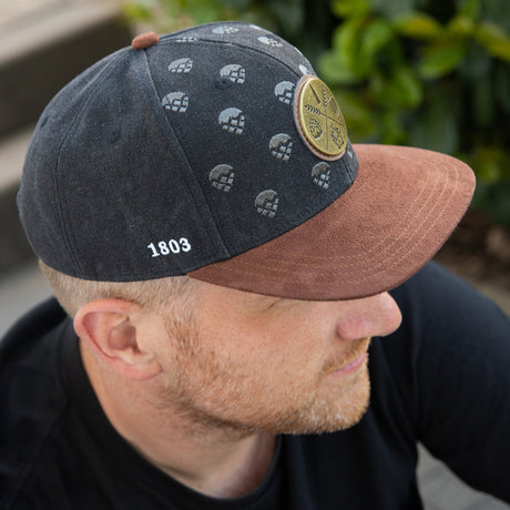 Stylische Krombacher Snapback in schwarz mit braunem Schild getragen von einem Mann der auf einer Treppe sitzt.