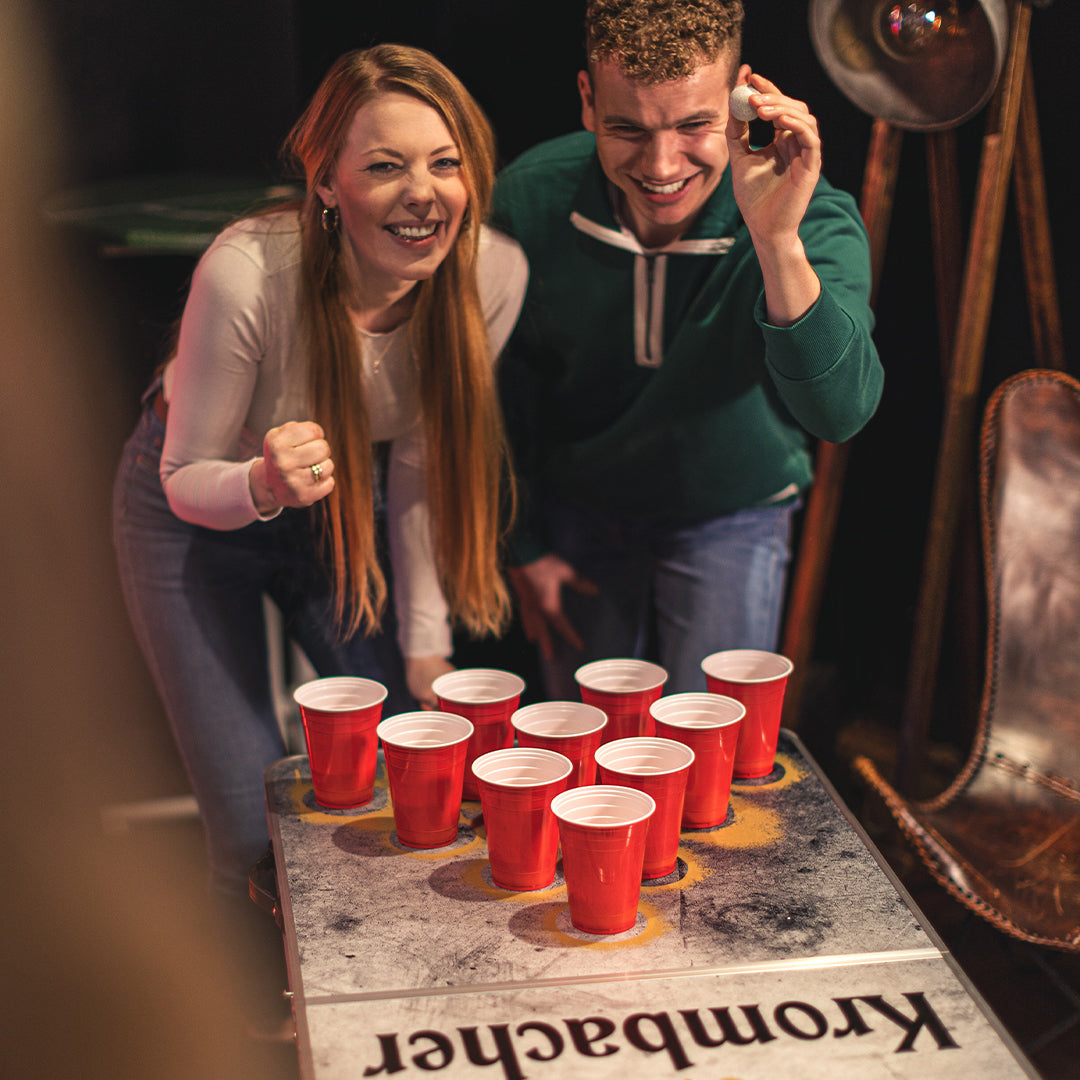 Zwei Personen spielen Beer Pong an einem Tisch mit roten Bechern, der Tisch trägt die Aufschrift 'Krombacher'.
