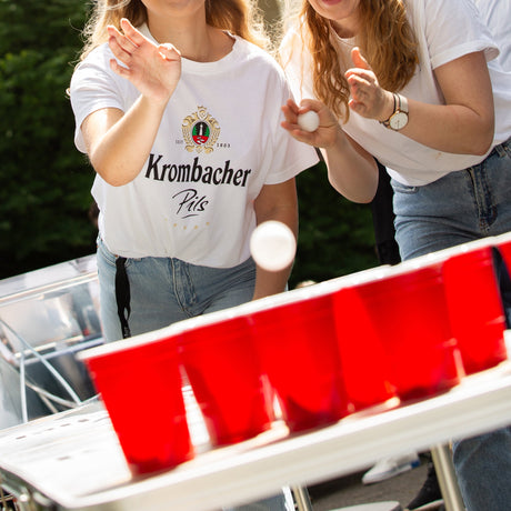 Zwei Frauen spielen Beer Pong, eine wirft einen Ball auf rote Becher, eine trägt ein weißes Krombacher Pils T-Shirt.