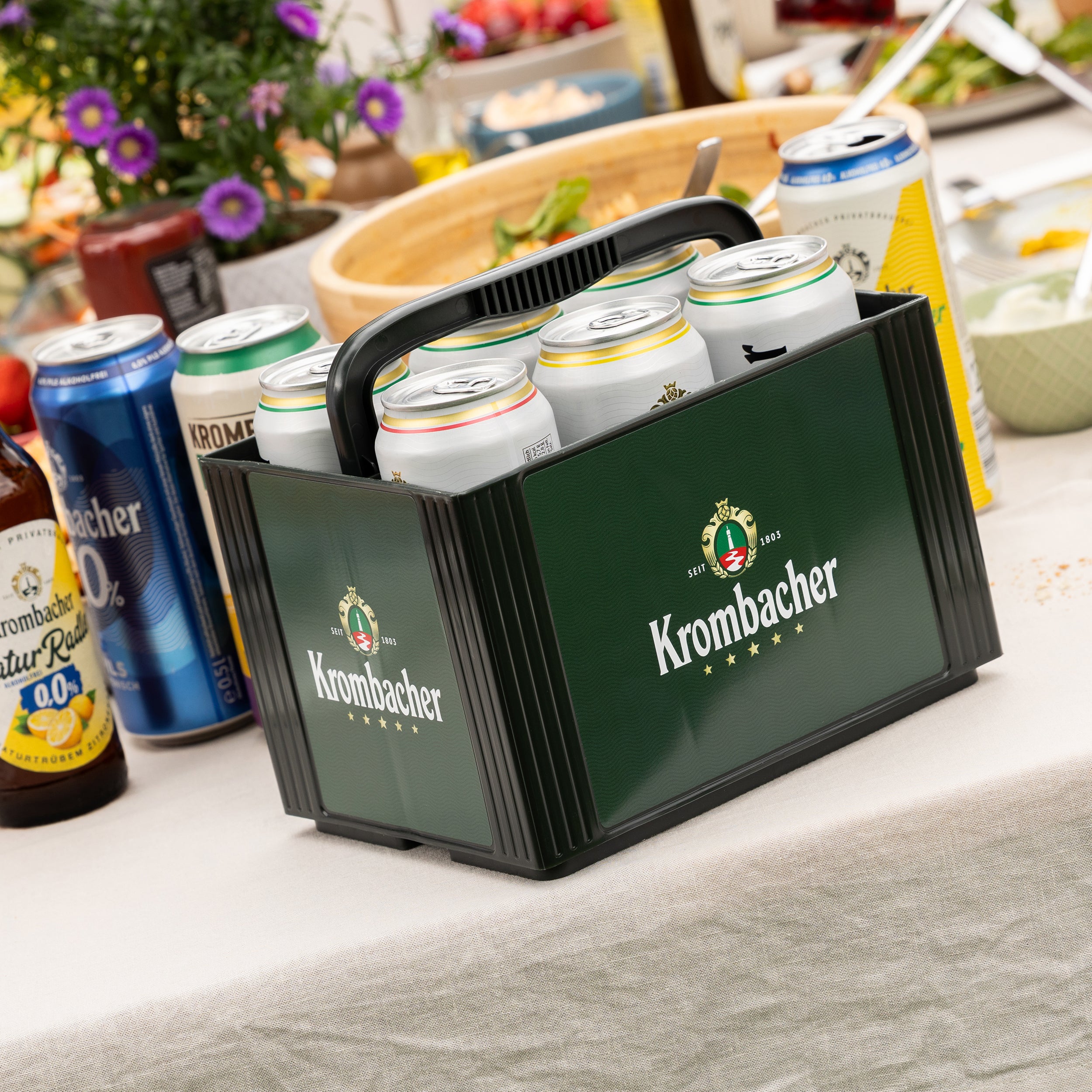 Sechserträger mit Krombacher-Dosen auf einem gedeckten Tisch mit weiteren Getränkedosen und Schalen im Hintergrund