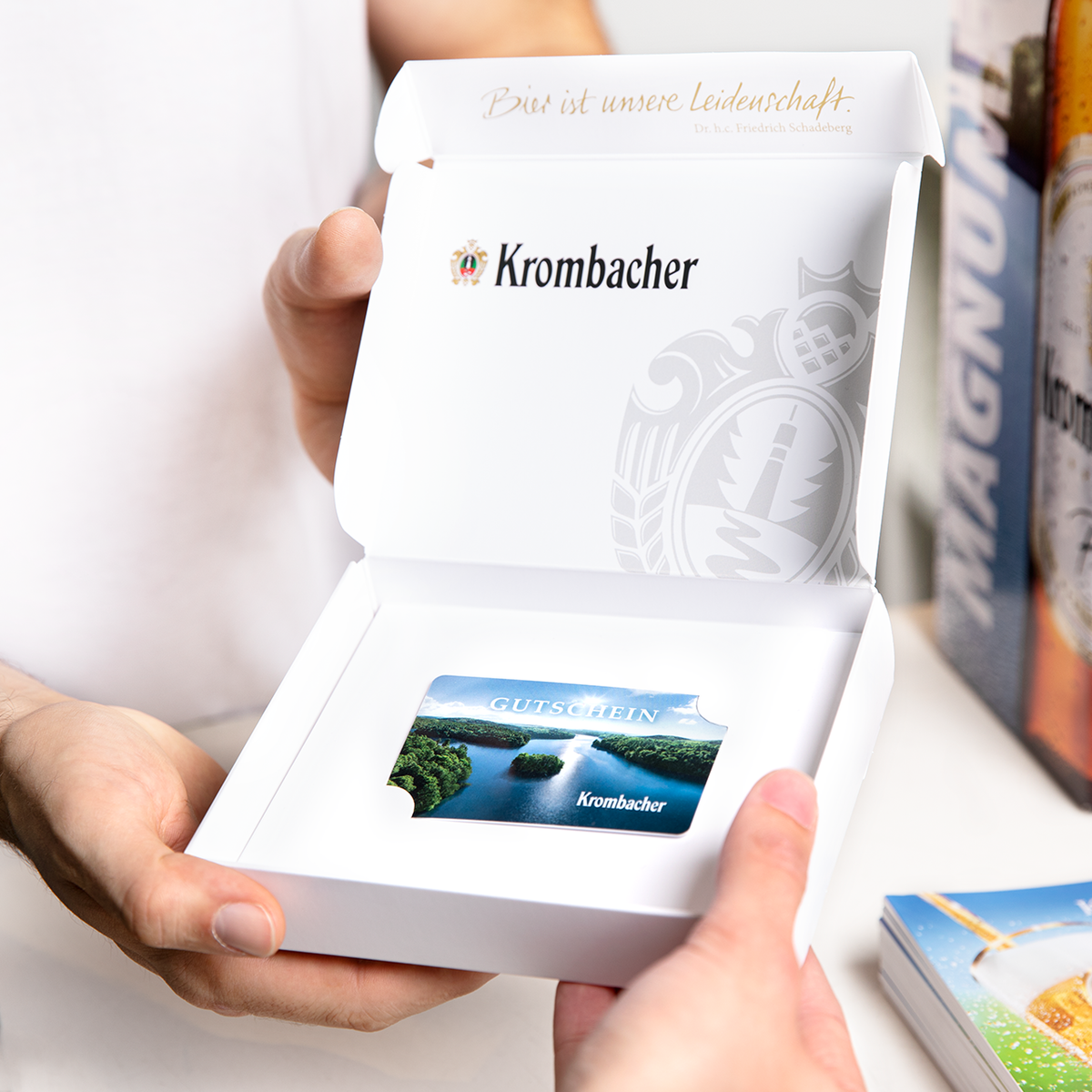 Person hält geöffnete weiße Krombacher-Gutscheinbox mit Gutscheinkarte und Aufschrift 'Bier ist unsere Leidenschaft'.