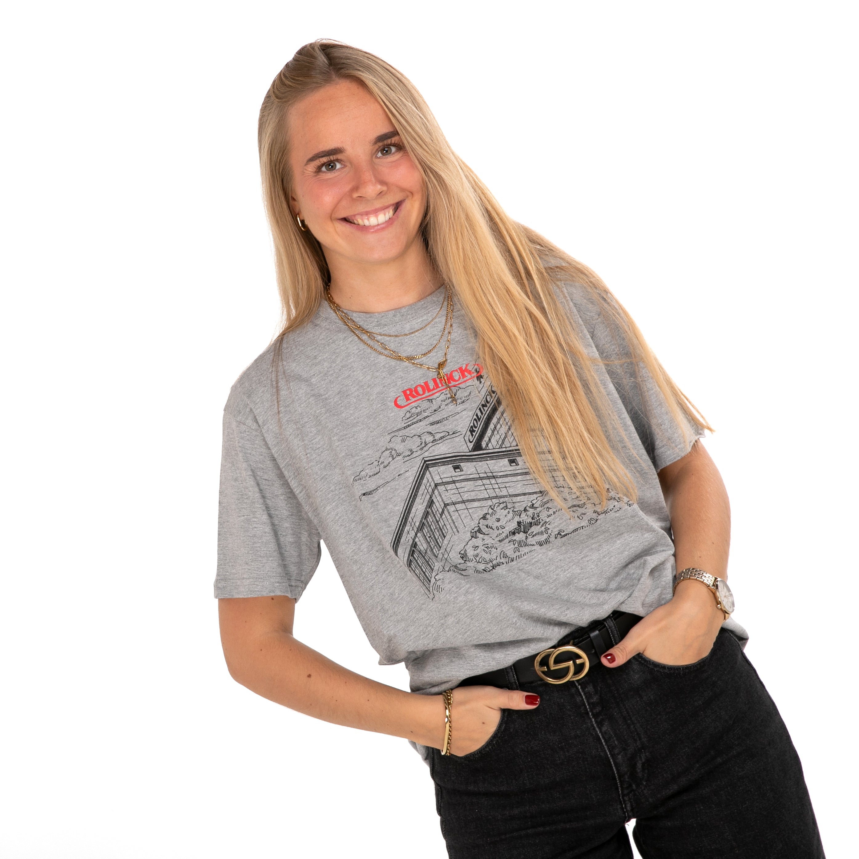 Frau mit langem blonden Haar trägt graues T-Shirt mit Rolinck Brauerei und rotem Schriftzug, schwarze Hose und goldene Accessoires.