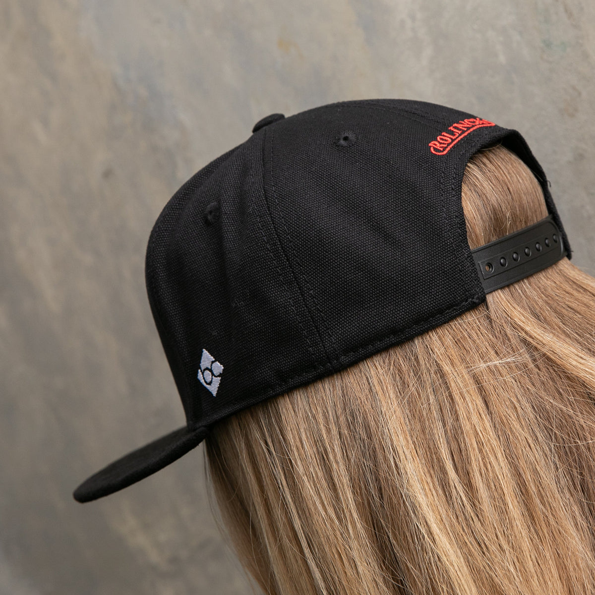 Schwarze Snapback-Cap mit roter Stickerei und verstellbarem Verschluss, getragen von hinten mit blondem Haar.