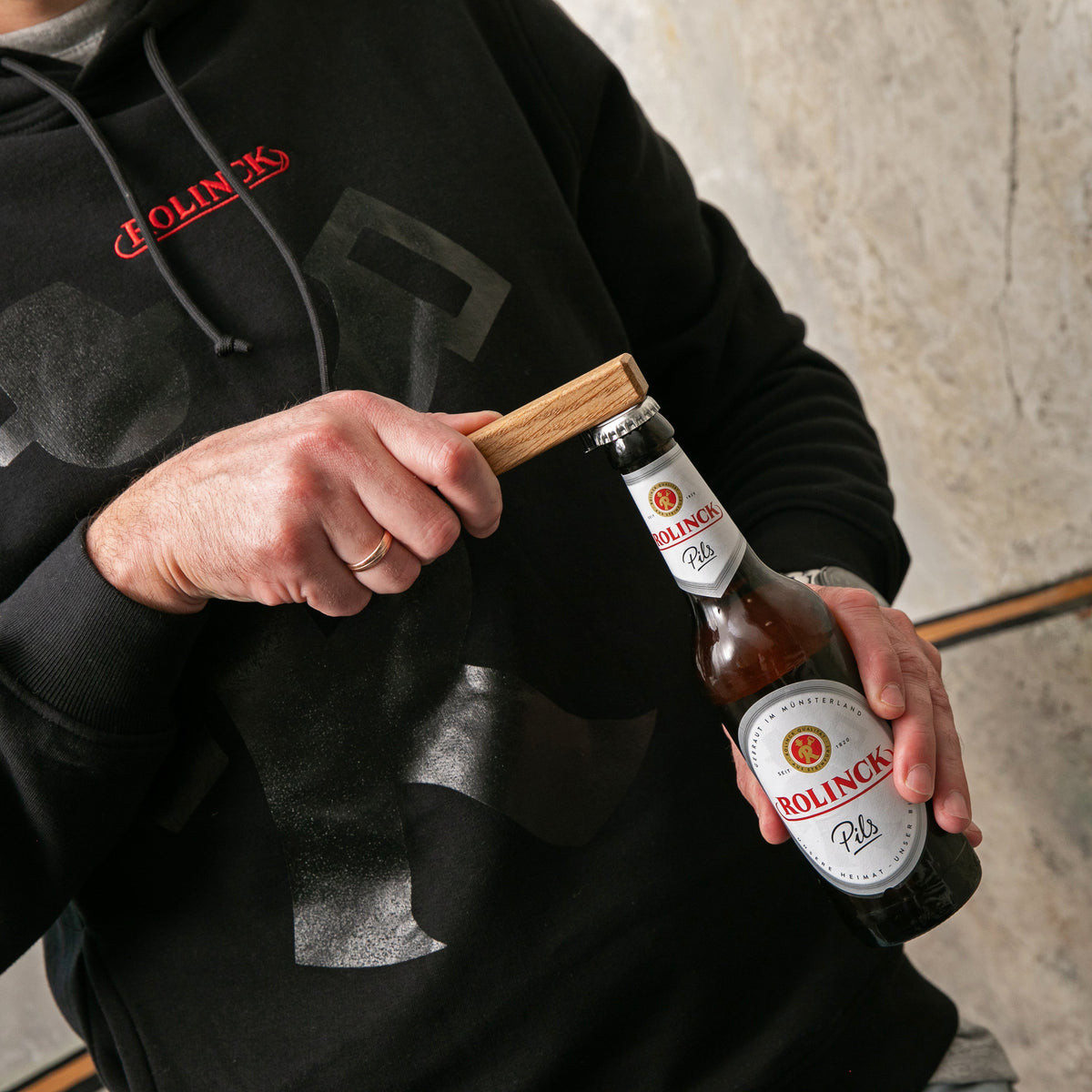 Person in schwarzem Hoodie mit rotem Schriftzug hält eine Flasche Rolnick Pils und öffnet sie mit einem Holz-Flaschenöffner.