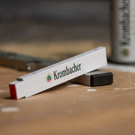 Zusammengeklappter weißer Zollstock mit grün-rotem Krombacher-Logo auf einer Werkbank