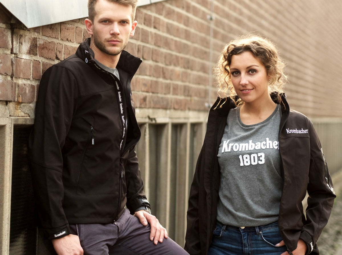 Zwei Personen lehnen an einer Backsteinwand, beide tragen schwarze Softshelljacken mit weißem Schriftzug 'Krombacher', die Frau trägt ein graues T-Shirt mit weißem Aufdruck 'Krombacher 1803'