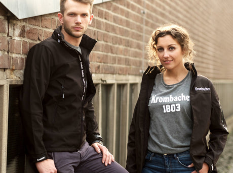 Zwei Personen lehnen an einer Backsteinwand, beide tragen schwarze Softshelljacken mit weißem Schriftzug 'Krombacher', die Frau trägt ein graues T-Shirt mit weißem Aufdruck 'Krombacher 1803'