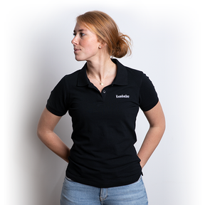 Frau trägt schwarzes Krombacher Damen Poloshirt mit kurzem Arm und weißem Logo auf der Brust, steht mit Händen in den Hüften.