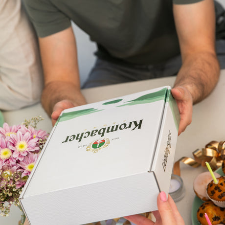 Zwei Personen übergeben sich eine weiße Krombacher-Verpackung mit grünem Design und Logo vor einem Tisch mit Blumen und dekorierten Muffins.