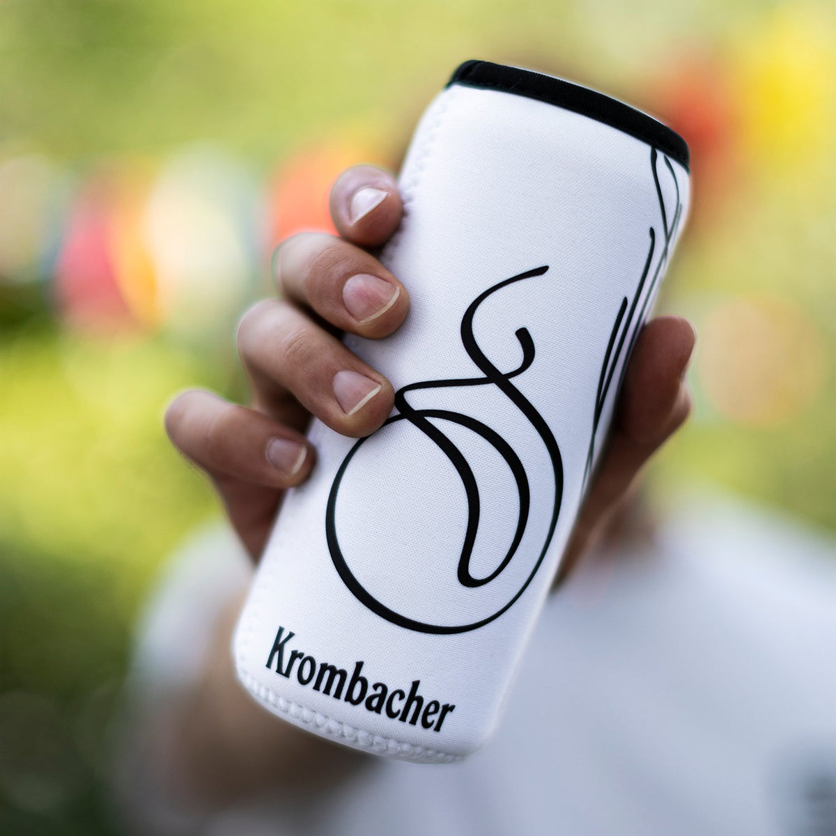 Hand hält weißen Dosenkühler mit schwarzer Krombacher-Logo-Schrift und geschwungenem Design.