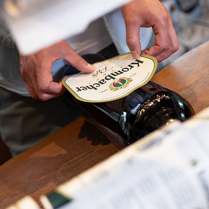 Krombacher Manufaktur: Ein Pils-Etikett wird von Hand auf eine Glasflasche geklebt.