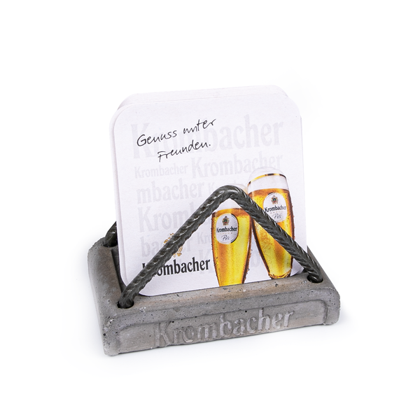 Bierdeckelhalter aus Beton mit geprägtem Schriftzug 'Krombacher' und zwei Bierdeckeln mit Krombacher-Logo und Text 'Genuss unter Freunden'.