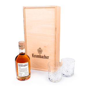 Geschenkset mit Krombacher Bierlikör in Flasche mit Holzverschluss und zwei Kristallgläsern vor einer Holzbox mit Krombacher-Logo.