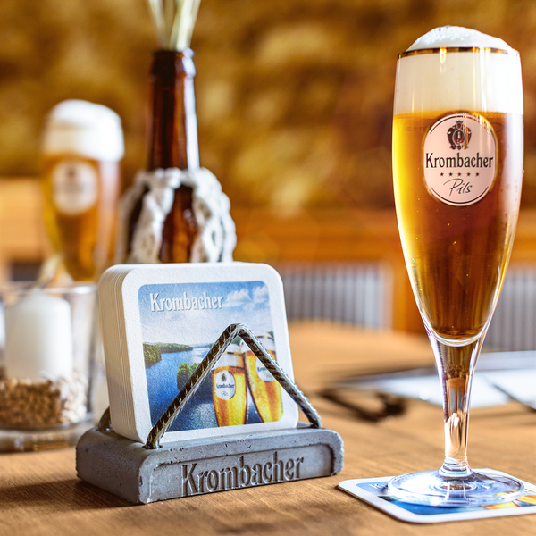 Krombacher Bierdeckelhalter aus Beton mit Bierdeckeln und ein Glas Krombacher Pils mit Schaum auf einem Holztisch.