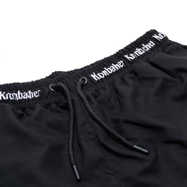 Schwarze Badehose mit elastischem Bund und weißem Schriftzug 'Krombacher' am Bund, mit schwarzem Kordelzug.