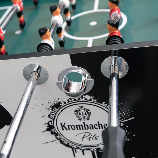 Krombacher Pils Kickertisch mit Spielfiguren in Rot und Weiß auf grünem Spielfeld, Metallstangen und Ballauswurföffnung.