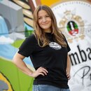 Eine Frau in einem schwarzen T-Shirt mit Illustriertem Kronkorken auf der Brust, vor einer bunten Wand.