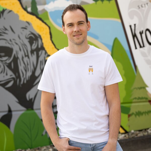 Ein Mann in einem weißen T-Shirt mit zwei anstoßenden Krombacher Gläsern auf der Brust, vor einer bunten Wand.