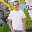 Ein Mann in einem weißen T-Shirt mit zwei anstoßenden Krombacher Gläsern auf der Brust, vor einer bunten Wand.