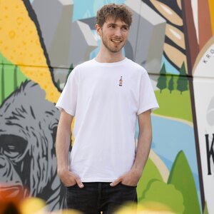 Ein Mann in einem weißen T-Shirt mit Krombacher Pils Flasche auf der Brust, vor einer bunten Wand.