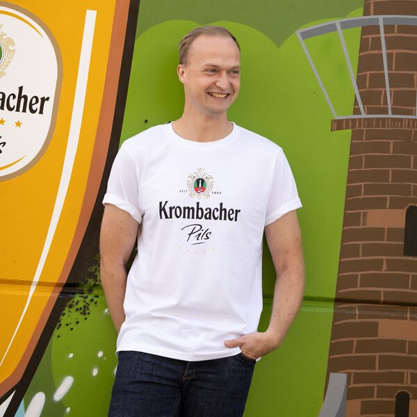 Ein Mann in einem weißen T-Shirt mit Krombacher Pils Logo auf der Brust, vor einer bunten Wand.