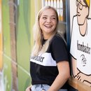 Eine Frau in einem schwarzen T-Shirt mit Krombacher Logo auf der Brust, vor einer bunten Wand.