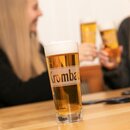 Krombacher Pils im Highlight Cup auf Holztisch, im Hintergrund zwei Personen die anstoßen.
